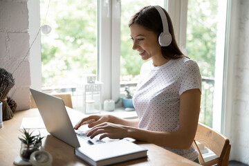 Happy millennial female sitting at desk in home office studying working online using laptop earphones. Positive young lady wearing headphones communicating in virtual web conference via social media