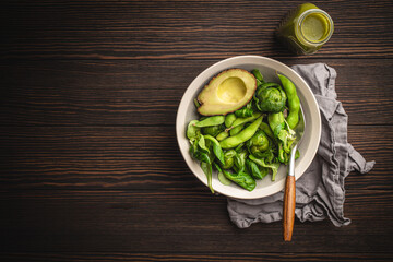 Green healthy salad with spinach, brussels sprouts, avocado in bowl and green detox smoothie in a bottle on wooden background, top view. Raw food diet and clean vegan eating, space for text
