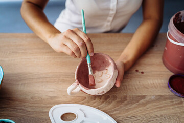 Close-up of girl painting clay mug. Woman coloring pottery in workshop with a paintbrush. Painter...