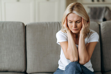 Middle aged blonde woman sits on couch at living room holding her head with her hands, feels unhappy because of headache, personal troubles, illness or bad news, she need psychological or medical