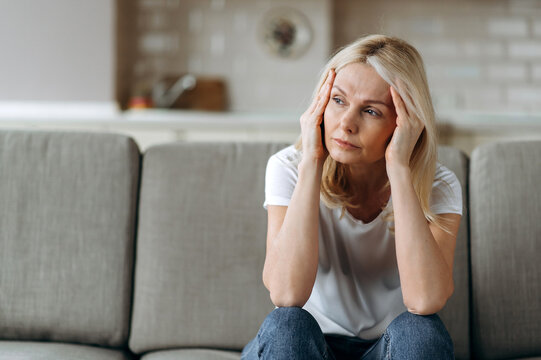 Sad caucasian senior woman wearing casual clothes sits on couch at home alone feels unhappy because of headache, stress, illness or bad news, she needs rest and sleep