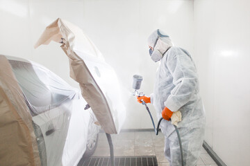 Car painter spraying white paint with spray gun on vehicle in paint chamber.
