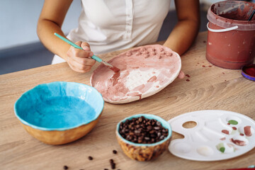 Close-up of hands. Selective focus. A young woman artist painting her clay product, a plate, in brown. Preparing for final burning. Brush on a colorless plate to make it bright and colorful later