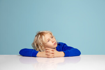 Attented. Happy curly boy isolated on blue studio background. Looks happy, cheerful. Copyspace for ad. Childhood, education, emotions, facial expression concept. Peeking out from behind the table