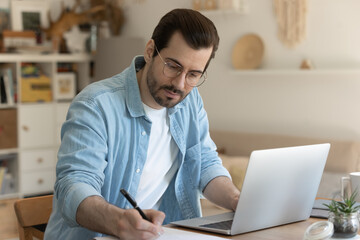 Diligent student. Concentrated millennial man studying online sit by laptop computer at home office...