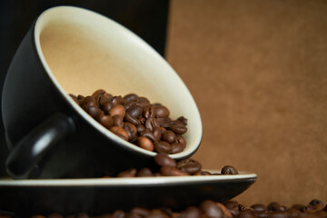 Ceramic cup filled with roasted black coffee beans, photographed on black wooden boards with paper bag and scattered coffee beans on the dark table.