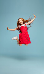 Weightless, freedom. Happy redhair girl in red dress on blue studio background. Looks happy, cheerful. Copyspace for ad. Childhood, education, emotions, facial expression concept. Jumping high, flying