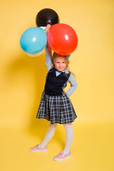 A girl in a school uniform with a festive cap on her head is holding balloons in her hands isolated on a yellow background.
