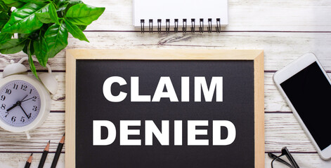 CLAIM DENIED written on a black background near pencils, a smartphone, a white notepad and a green plant in a pot