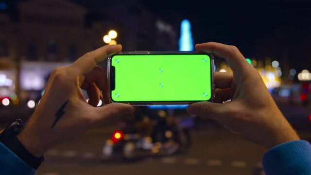 Man hold smartphone with green screen. Background of busy bustling night city life, traffic, light and cars. Smartphone perfect for product placement and integration. Man use phone for application