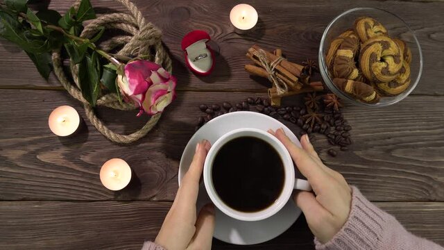 Female hands on a white cup of coffee. Table decoration for Valentine's Day.A romantically decorated table with candles, a rose flower and a gold ring in a box.