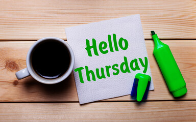 On a wooden table, a cup of coffee, a green marker and a napkin with the text HELLO THURSDAY