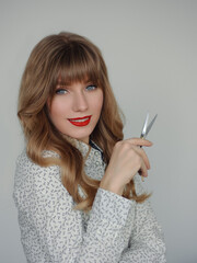 Obraz premium Attractive young blonde makeup artist woman in white sweater holding professional hair scissors posing for blog in social networks enjoying beauty routine