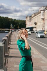 young beautiful blonde woman with red lipstick and in a green dress talking on a mobile phone in the city on the embankment of the river