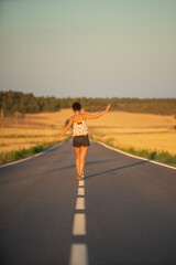 Obraz premium Young girl, walking in the middle of a lonely straight road, late in the day. With the hands stretched out in the shape of a cross, to maintain balance and not fall from the dividing line.