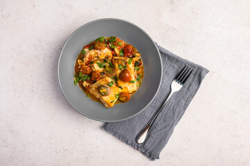 Traditional Sicilian fish with tomatoes, olives, parsley and red pepper in a deep plate with napkin and fork on a light background, close up