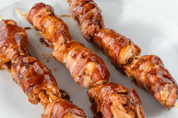 Grilled bacon-wrapped chicken tenders are laid out on a white plate. Strung on wooden skewers.