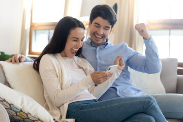Overjoyed young man and woman using phone, celebrating success, showing yes gesture, rejoicing...