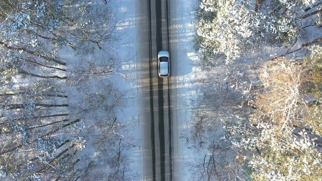 Aerial shot of white car riding through snow covered icy road. SUV driving at empty countryside route in the forest on winter day. Flying over the auto moving through scenic landscape way. Top view