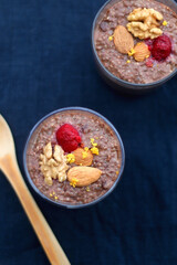 Two glasses of chocolate, orange and chia seed pudding with nut and raspberry topping. Top view, dark blue background.