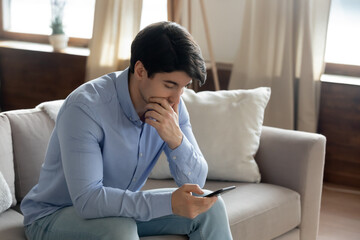 Close up thoughtful serious man touching chin, looking at smartphone screen, having problem with phone or reading bad news in message, unexpected debt notification, upset male sitting on couch