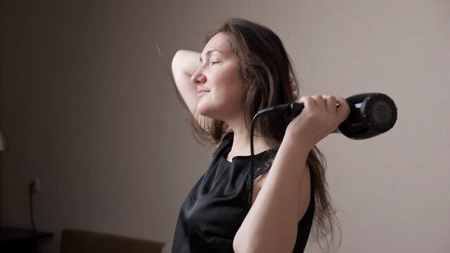 Nice and calm lady in black t-shirt enjoys drying long hair with modern styler standing against white wall in room close view slow motion