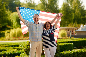 Obraz premium Man and woman holding the usa flag. Patriotic married mature couple. Green summer park background.
