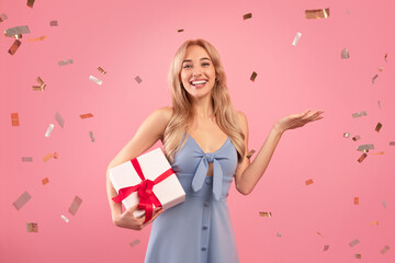 Joyful young lady in lovely dress holding wrapped gift box on pink studio background with falling...