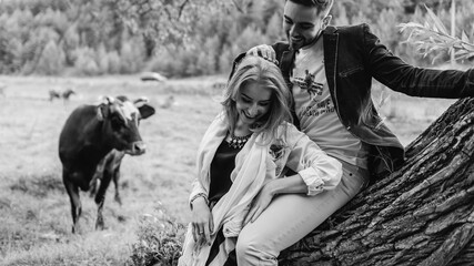 Fototapeta na wymiar Handsome guy and blonde girl walking on the grass near the river and forest. They hold hands and smile on the background of pasture with cows and horses