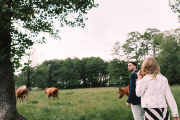 Handsome guy and blonde girl walking on the grass near the river and forest. They hold hands and smile on the background of pasture with cows and horses