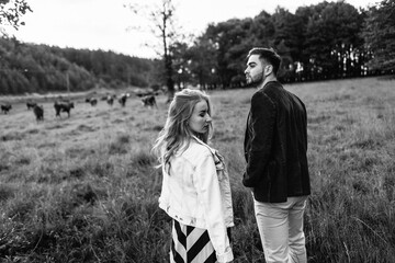 Handsome guy and blonde girl walking on the grass near the river and forest. They hold hands and smile on the background of pasture with cows and horses