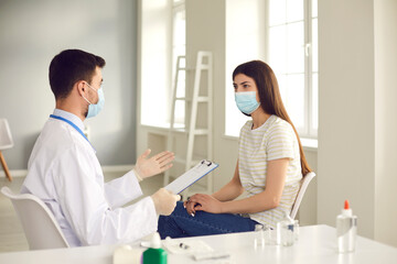 Male doctor talking to female patient during vaccination campaign at clinic or hospital. Medical worker in face mask interviewing young woman before giving her Covid-19 vaccine. Concept of health care
