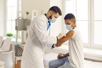 Doctor in medical face mask disinfects skin on patient's arm before injecting him with flu vaccine. Young African-American man getting Covid-19 antiviral shot during vaccination campaign at the clinic - obrazy, fototapety, plakaty