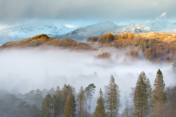 Beautiful golden light illuminating Cumbrian fells on a Winter afternoon with mist filled valley and snowcapped mountains. Lake District, England, UK.
