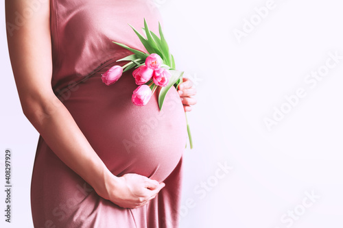 Beautiful pregnant woman with tulips flowers holds hands on belly. Young woman in dress waiting for baby birth. Pregnancy, Motherhood, Mother's Day Holiday concept.