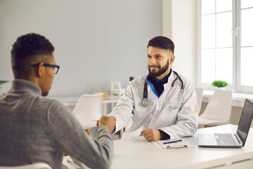 Obraz premium Smiling man doctor and african american man patient shaking hands after successful checkup consultation in medical clinic office. Healthcare, medicare, communication with doctor concept