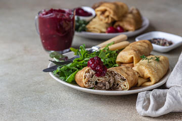 Homemade stuffed thin pancakes or crepes with meat, spicy cranberry sauce and arugula, light stone background. Traditional Russian cuisine for Maslenitsa. Shrove Tuesday.