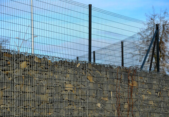 retaining wall made of gabon baskets. there is still a wire fence on top of a high gray wall. under...