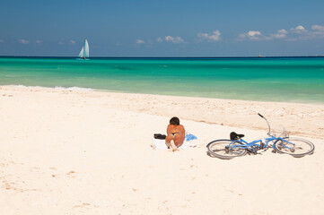 Fototapeta na wymiar Young Caucasian woman unrecognizable, Tanning on a deserted Caribbean beach with his bicycle on the sand. On the horizon, the blue sky and a sailboat, Playa del Carmen, Mexico.