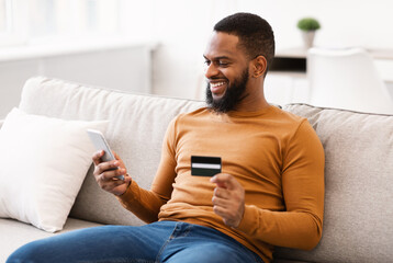 African Guy Using Credit Card And Smartphone Sitting At Home