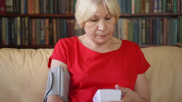 Senior woman measure pressure by tonometer at home during lockdown. Female pensioner sick during pandemic. Bookcase bookshelves in the background. Concept of social distancing