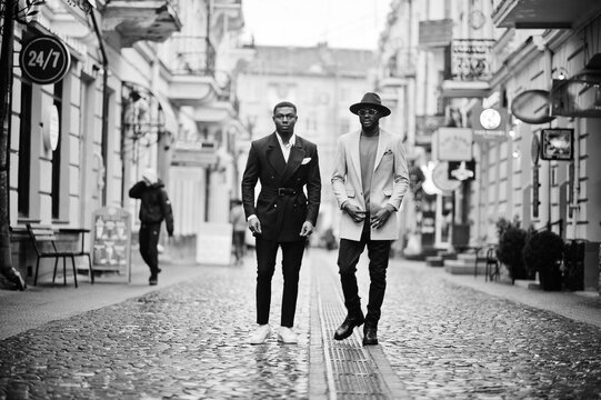 Two fashion black men walking on street. Fashionable portrait of african american male models. Wear suit, coat and hat.