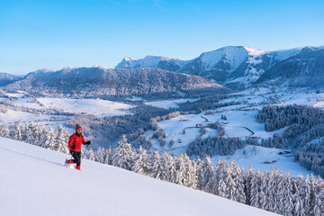 nice active senior woman snowshoeing in the Allgaeu Alps near Oberstaufen with view on the Nagelfluh mountain chain with Mount Hochgrat and village of Steibis, Bavaria, Germany