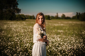 Fototapeta na wymiar Young fashionable woman holding bunch of daisies in her hands looking to camera and wearing a lace playsuit in the middle of daisy meadow
