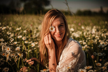 Simple portrait of young woman looking into camera, hiding one eye with daisy and sitting in a daisy meadow wearing white lace playsuit.