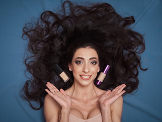 Fototapeta premium Skin care beautiful cosmetic makeup mixed race brunette model gorgeous hair full lips tan olive skin lying cobalt color backdrop professional cosmetic bottles and brushes