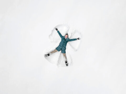 Young beautiful woman in a gray jacket, light pants, boots makes a snow angel in the snow, a view from a drone. Snow fun, entertainment.