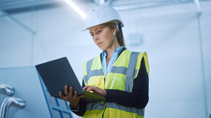 High-Tech Factory: Confident and Professional Female Engineer Wearing Safety Jacket and Hard Hat Holding and Working on Laptop Computer. Modern Bright Industrial Facility. Low Angle Shot