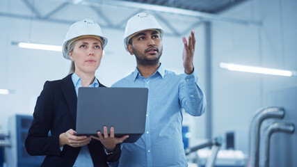 Female Chief Engineer and Male Project Managers Standing in the Middle of Modern Factory, Use Laptop Computer, Plan Production Line Optimization. Professional People work on Industrial CNC Machinery