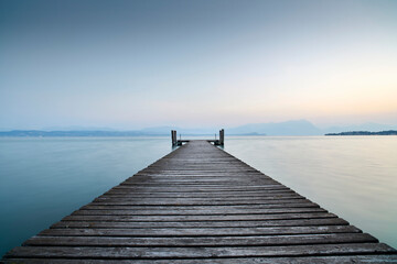 Fototapeta na wymiar An old wooden pier extends into the clear blue waters of the lake. A solitary path towards the calm and peaceful silence of nature, with the sweet sound of the waves.